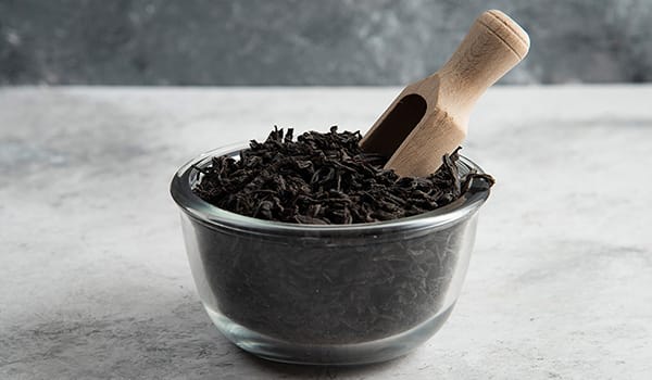 Glass bowl of loose black tea leaves with a wooden scoop resting in them on a light gray surface and countertop.